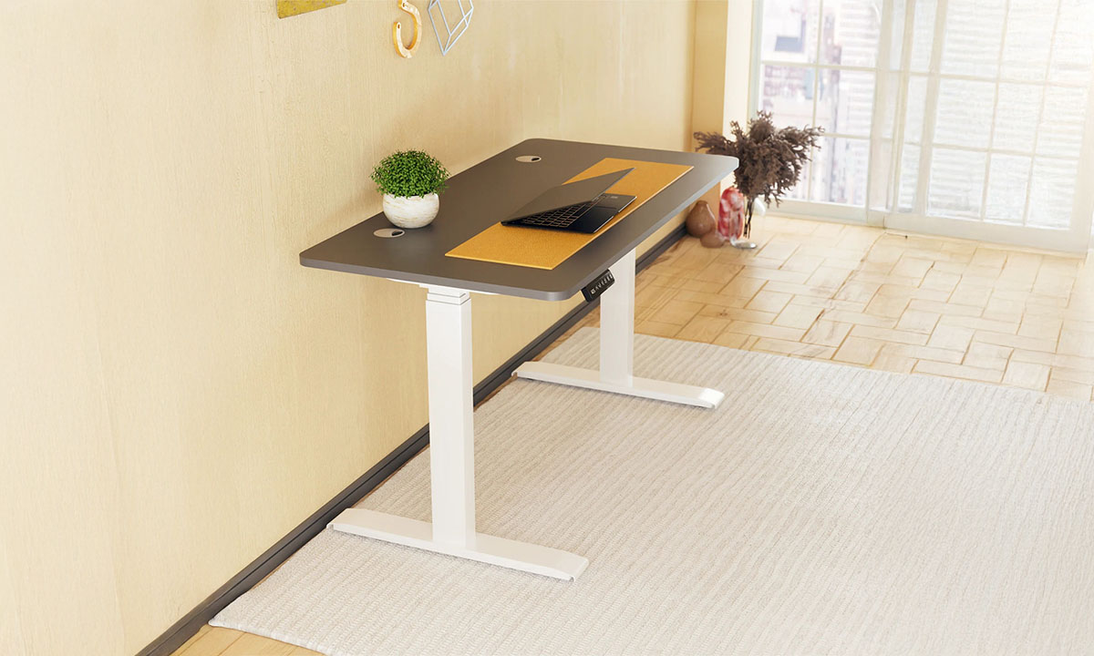 Modern çalışma masası, açık renk duvar ve büyük pencere ile aydınlatılmış ofis ortamında kullanılıyor. Çalışma alanında laptop, dekoratif objeler ve bitki bulunuyor.