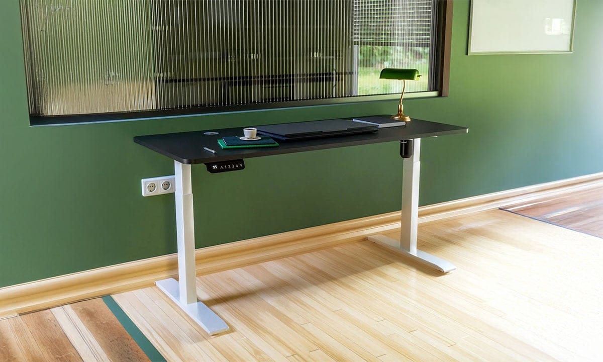 Yüksekliği ayarlanabilir iki motorlu ofis çalışma masası, modern siyah yüzeyli, yeşil duvar ve doğal ahşap zeminle şık bir çalışma ortamı.