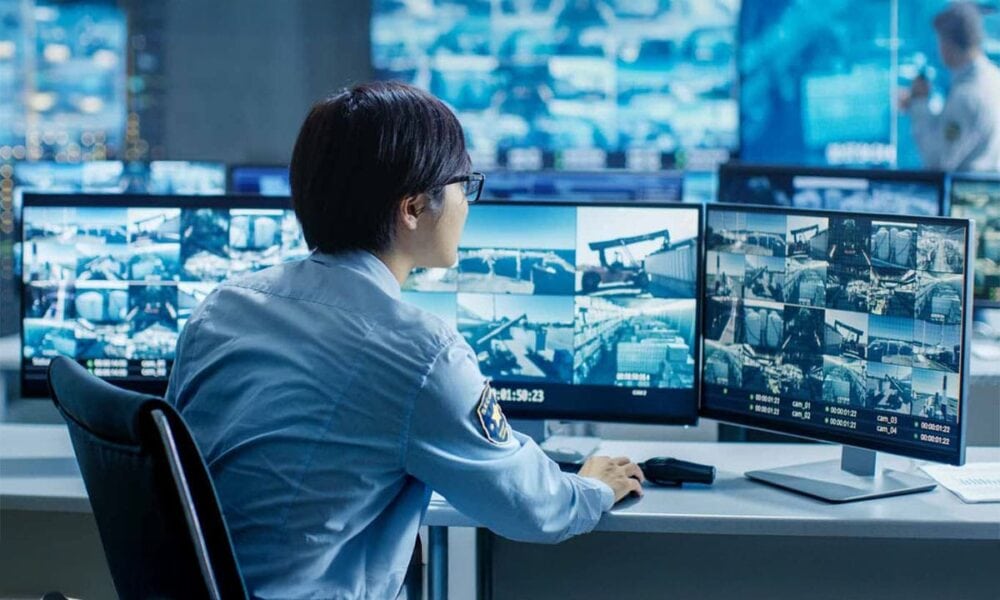 Yedekleme ve güvenlik görevlisi izleme merkezinde çoklu monitörler kullanılarak güvenlik kameraları ve sistemleri izleniyor. Bu yüksek teknolojili ortamda, güvenlik ve veri yönetimi ön planda tutuluyo.